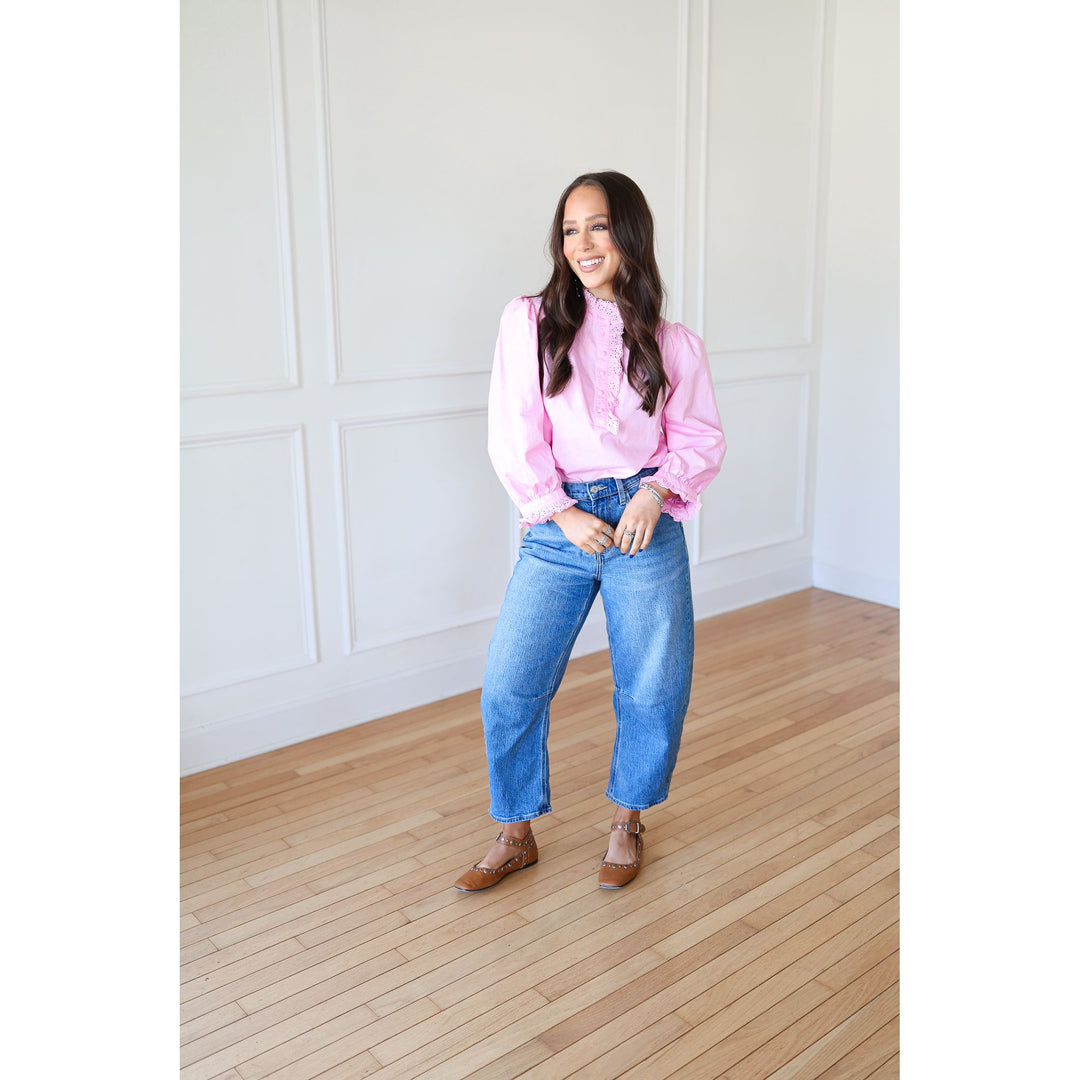 Woman wearing a pink blouse and blue jeans standing in a room with wooden flooring and white walls.