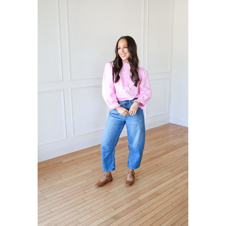 Woman wearing a pink blouse and blue jeans standing in a room with wooden flooring and white walls.