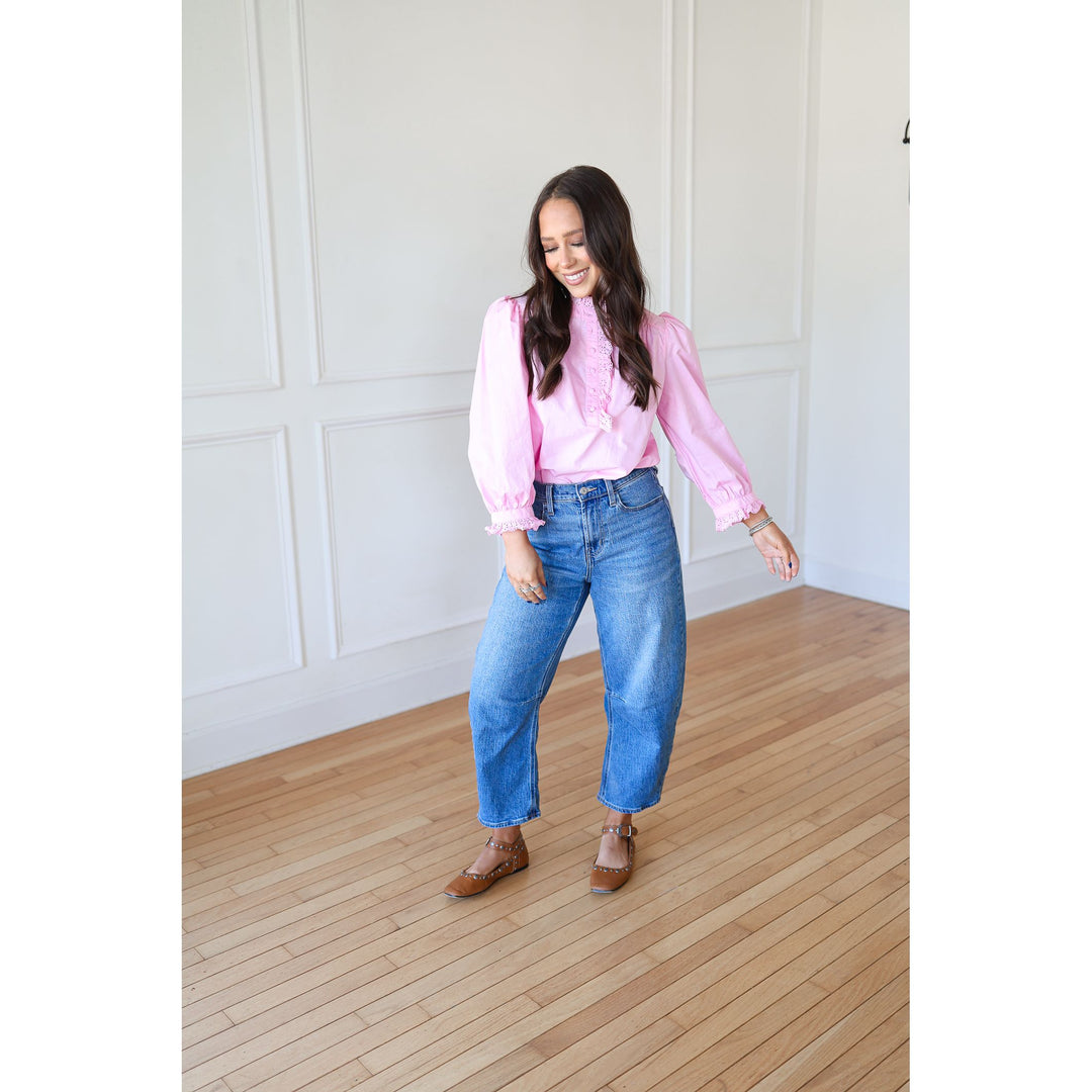 Woman wearing a pink blouse and blue jeans standing in a room with wooden flooring and white walls.