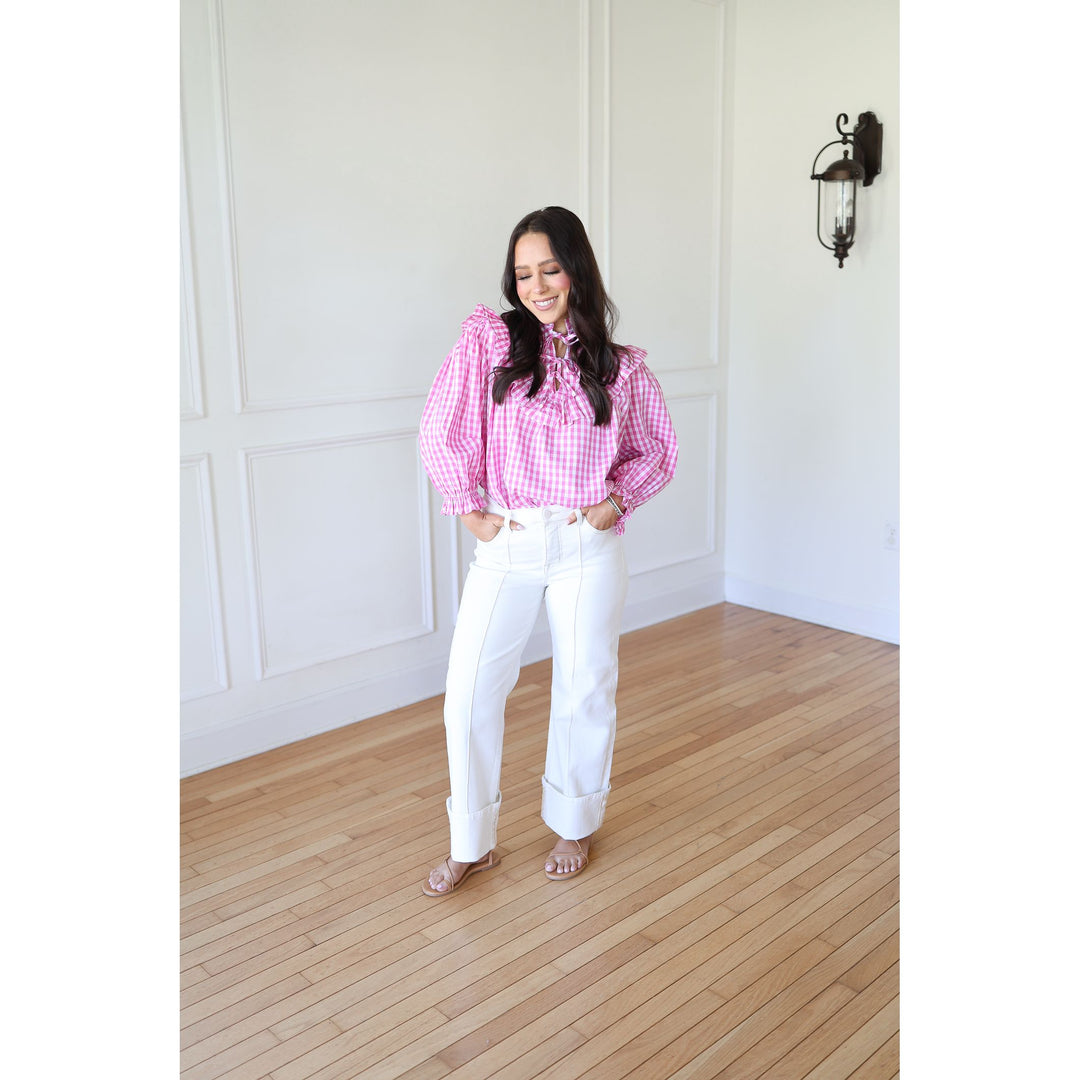Woman wearing a pink checkered blouse and white pants standing in a room with wooden flooring and white walls.