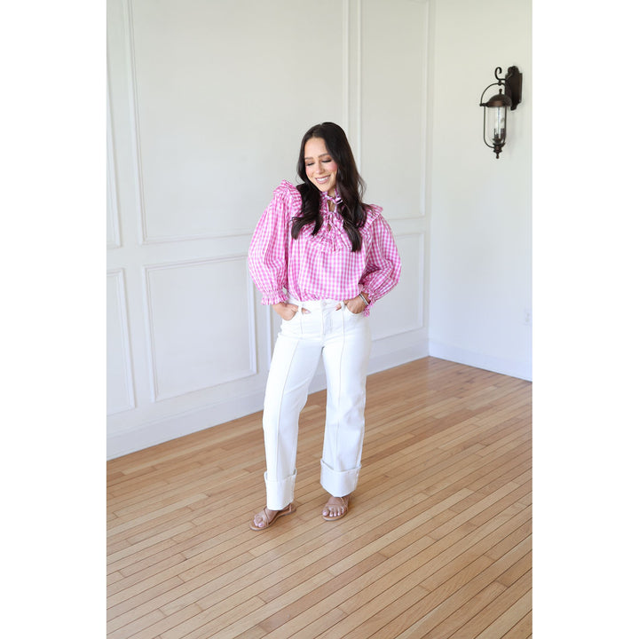 Woman wearing a pink checkered blouse and white pants standing in a room with wooden flooring and white walls.