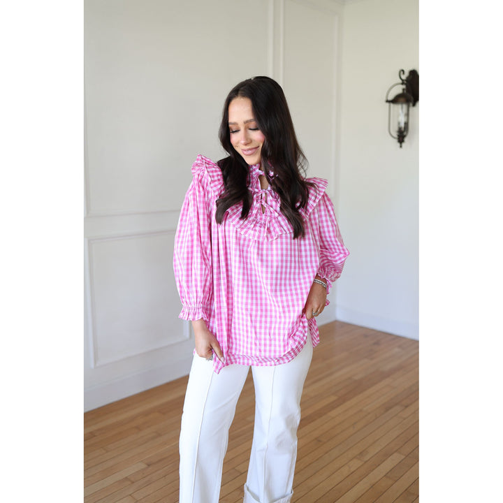 Woman wearing a pink checkered blouse and white pants in a room with wooden flooring and white walls.