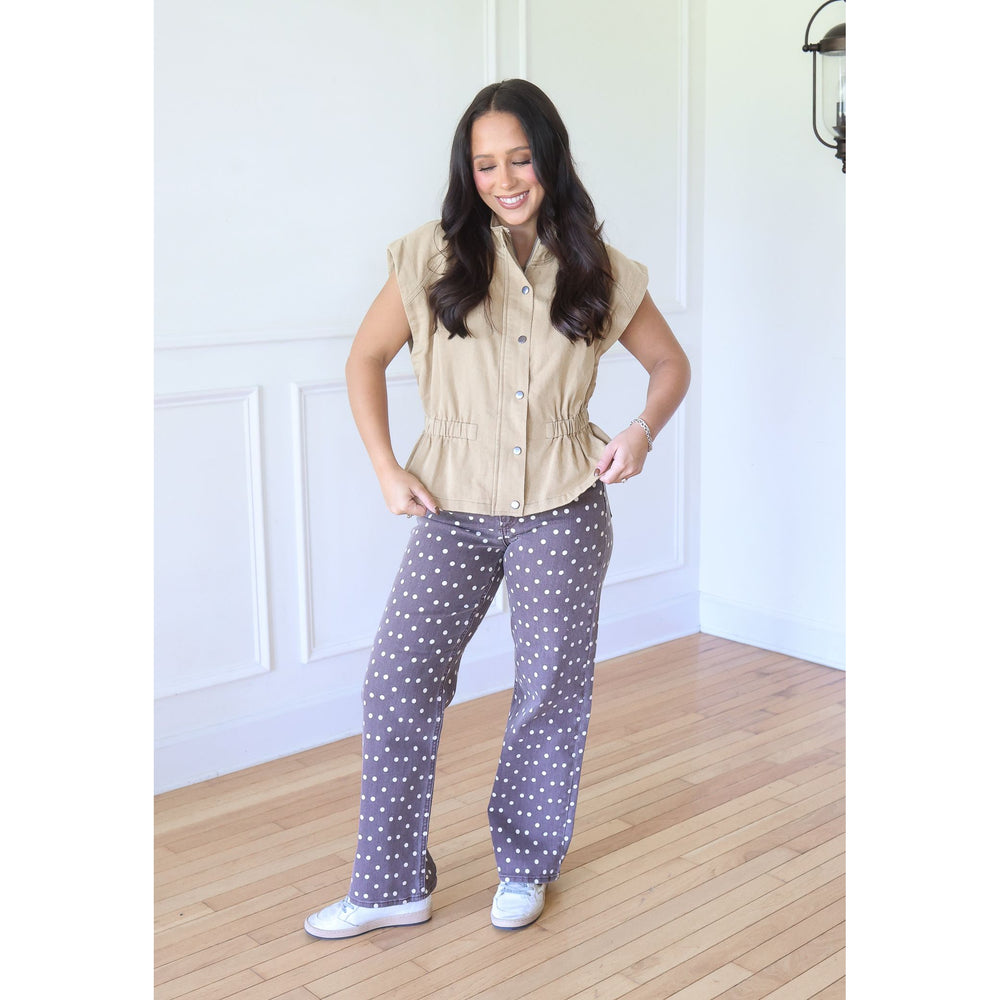 A woman modeling a beige sleeveless button-up shirt and gray pants with white polka dots, standing in a bright room with white paneled walls and light wood floors.