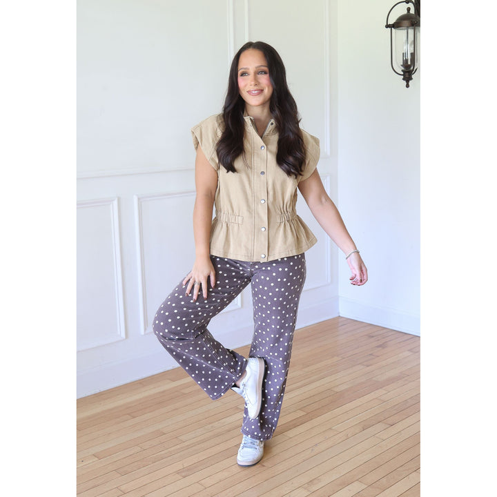 A woman modeling a two-piece pajama set featuring a beige short-sleeve button-up shirt and dark gray pants with white polka dots, standing on a light wood floor against a white paneled wall.