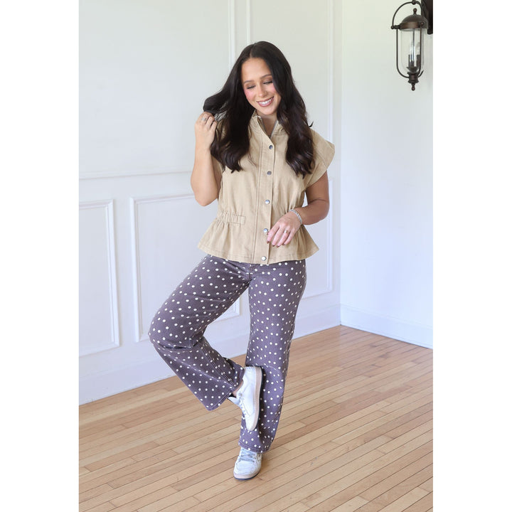 A woman modeling a two-piece pajama set in a bright room with white paneled walls and light wood floors. The set includes a beige short-sleeve button-front top and dark gray pants with white polka dots.