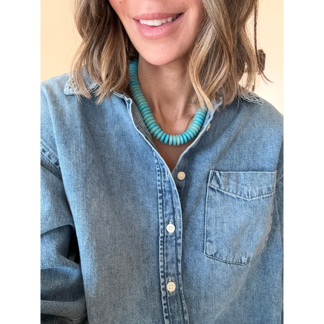 Person wearing a blue denim shirt and teal necklace against a neutral background