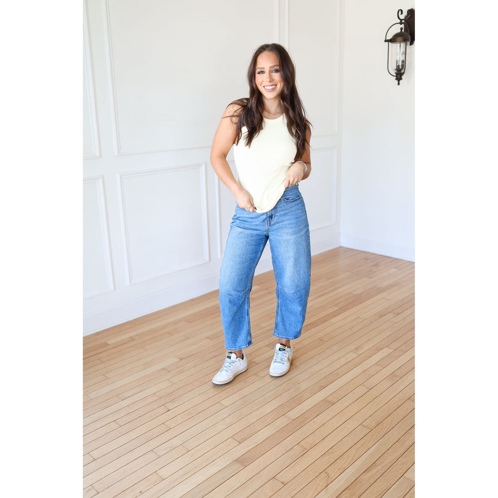 Woman wearing a yellow top and blue jeans standing in a room with wooden flooring and white walls.