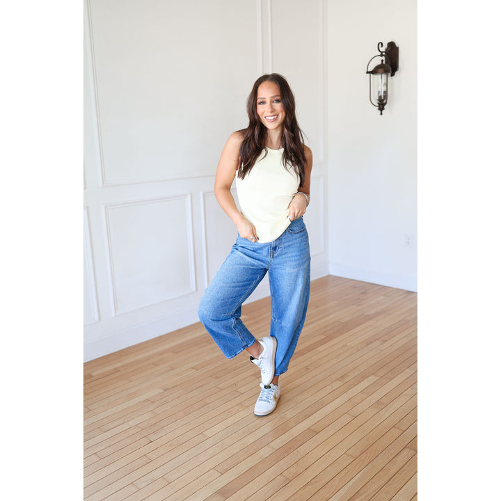 Woman wearing a white tank top and blue jeans standing on a wooden floor.