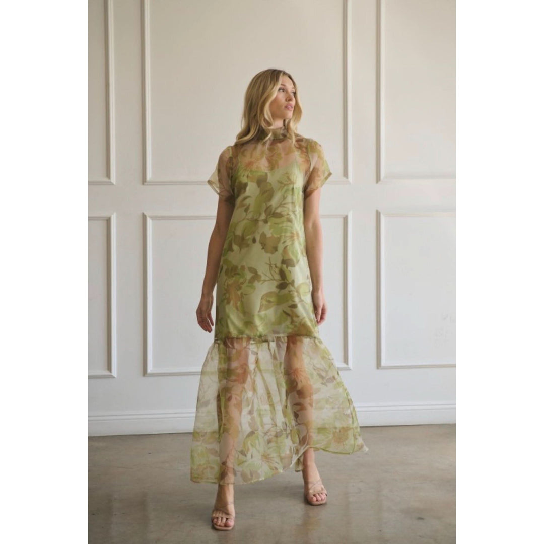 Woman wearing a green floral dress standing against a white paneled wall.