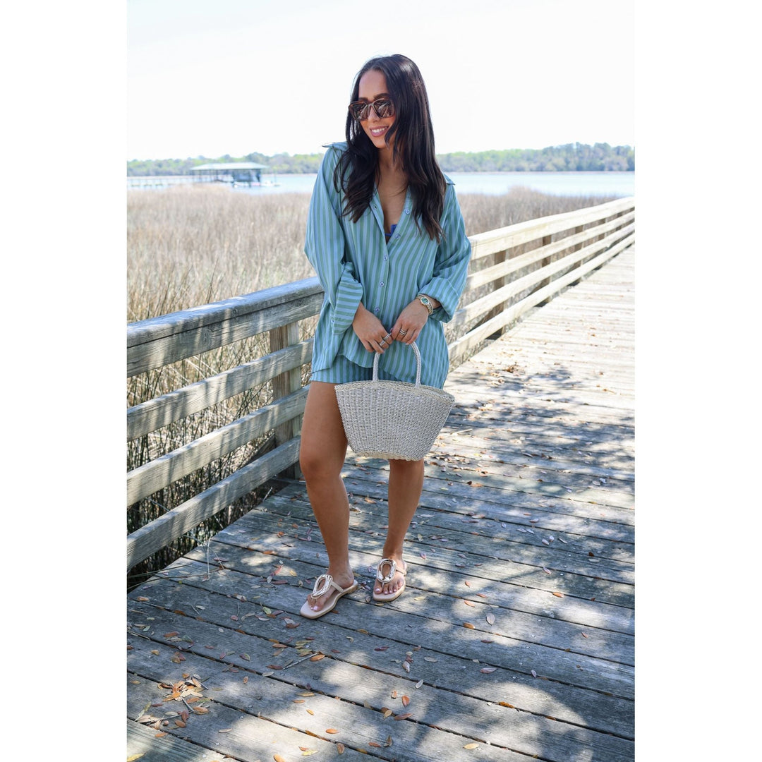 Woman in a blue striped shorts set holding a woven bag on a wooden boardwalk by a body of water.