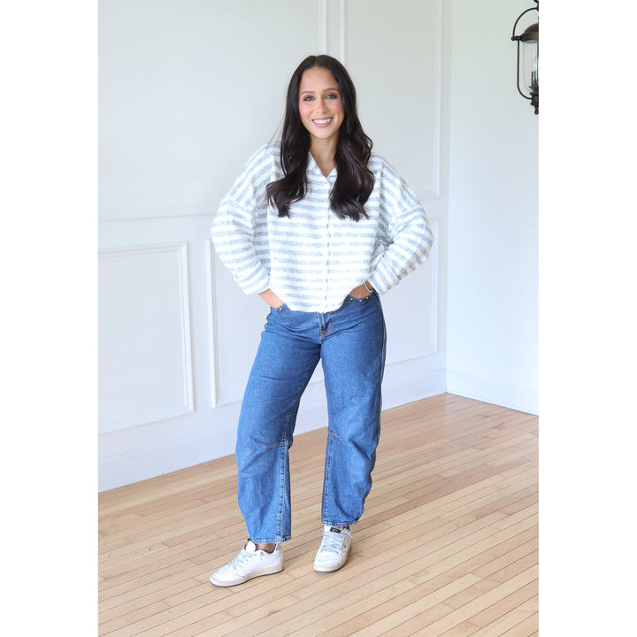 A woman stands in a bright room with white paneled walls and light wood floors, wearing a white and gray blue striped button-up shirt, blue jeans, and white sneakers.