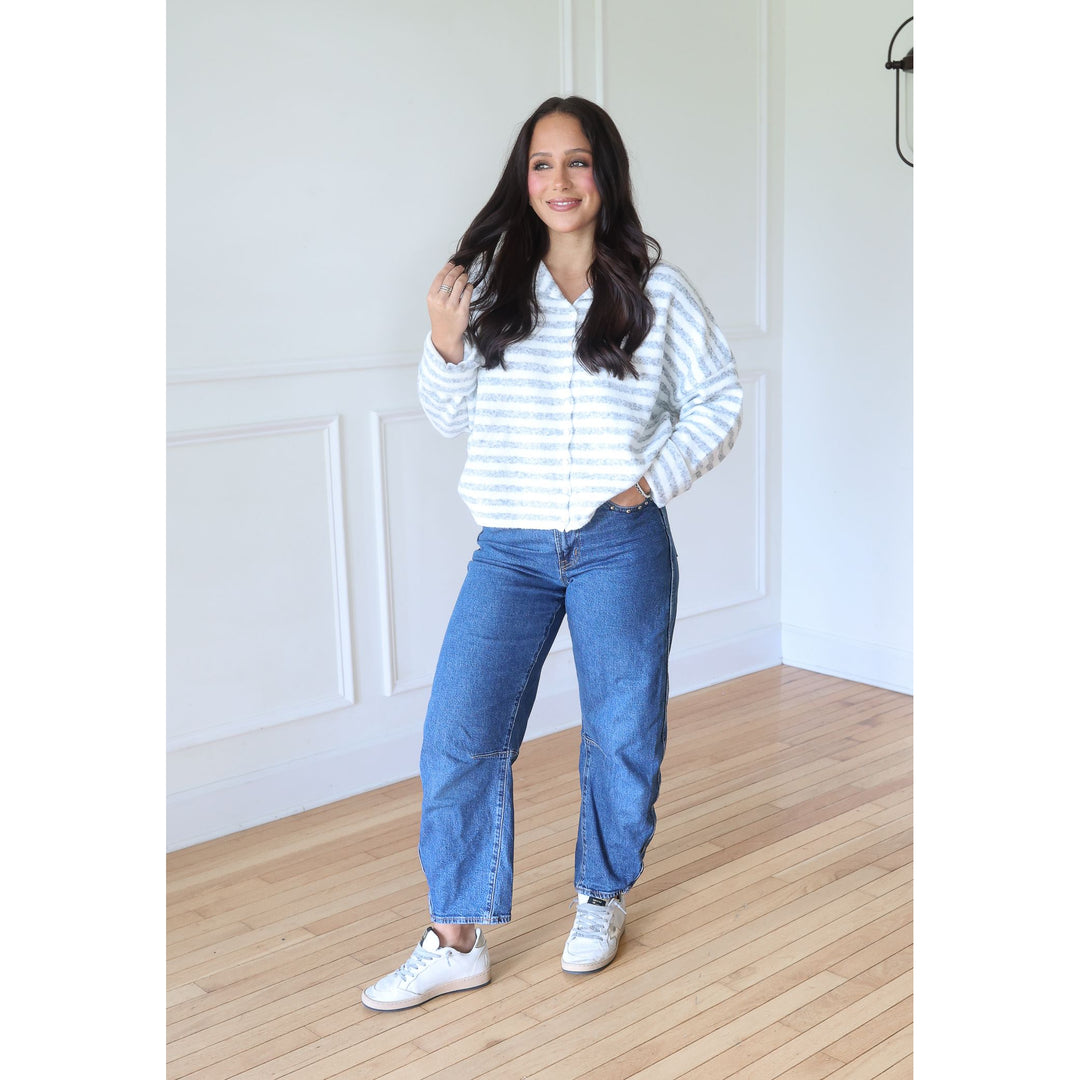A woman modeling a white and gray blue horizontally striped long-sleeve button-up shirt, paired with blue jeans and white sneakers, standing in a bright room with white paneled walls and light wood floors.