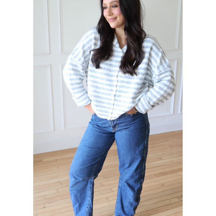 A woman wearing a white and light blue striped button-up shirt with blue jeans, standing in a bright room with white paneled walls and a light wood floor.