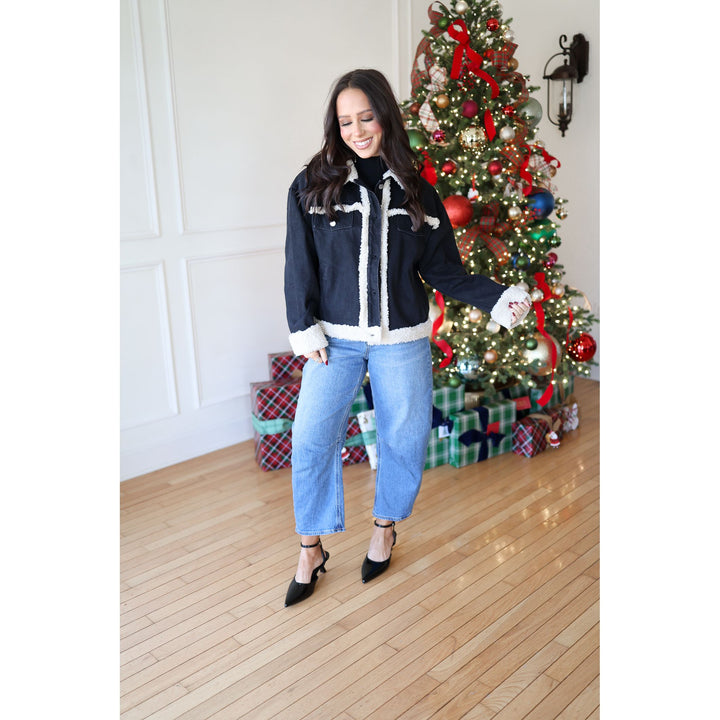 Woman in a black denim jacket and jeans standing in front of a decorated Christmas tree.