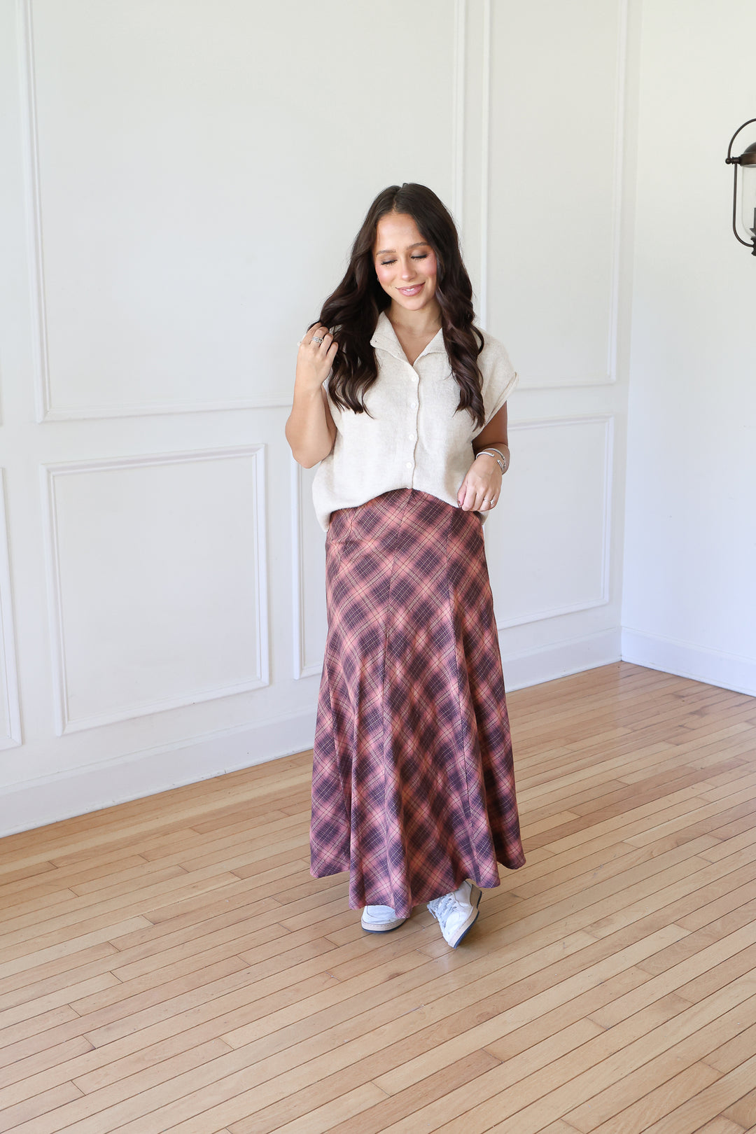 A woman modeling a long plaid skirt with a white sleeveless top, standing on a light wood floor against a white paneled wall.