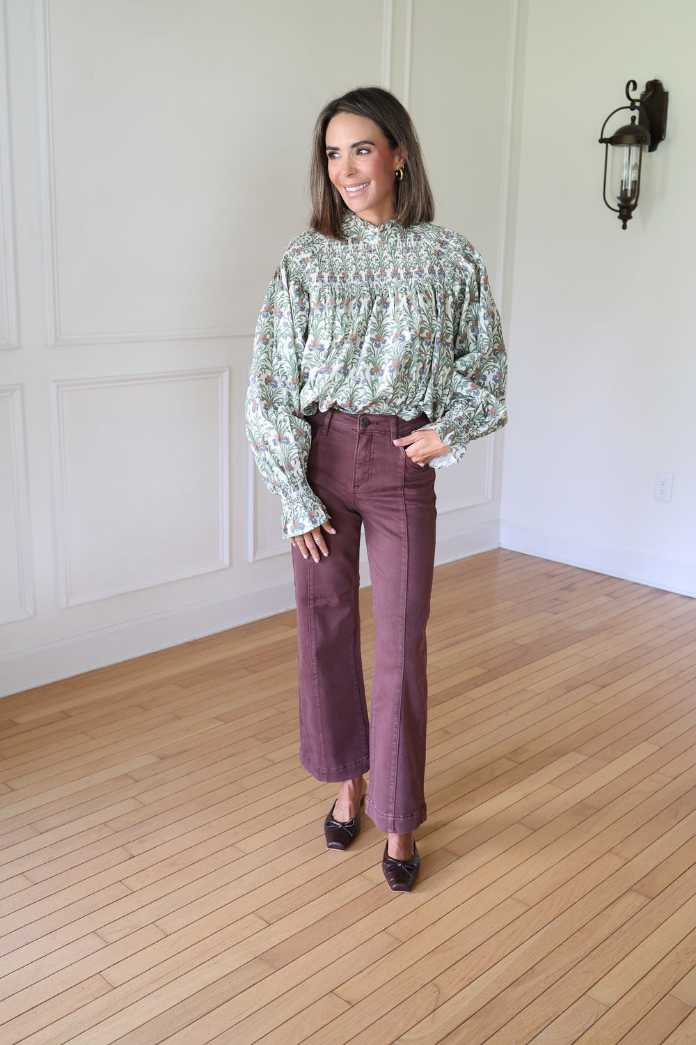 A woman stands in a bright room with white paneled walls and light wood floors, wearing a long-sleeve floral blouse with a ruffled high neck and dark mauve wide-leg cropped pants.
