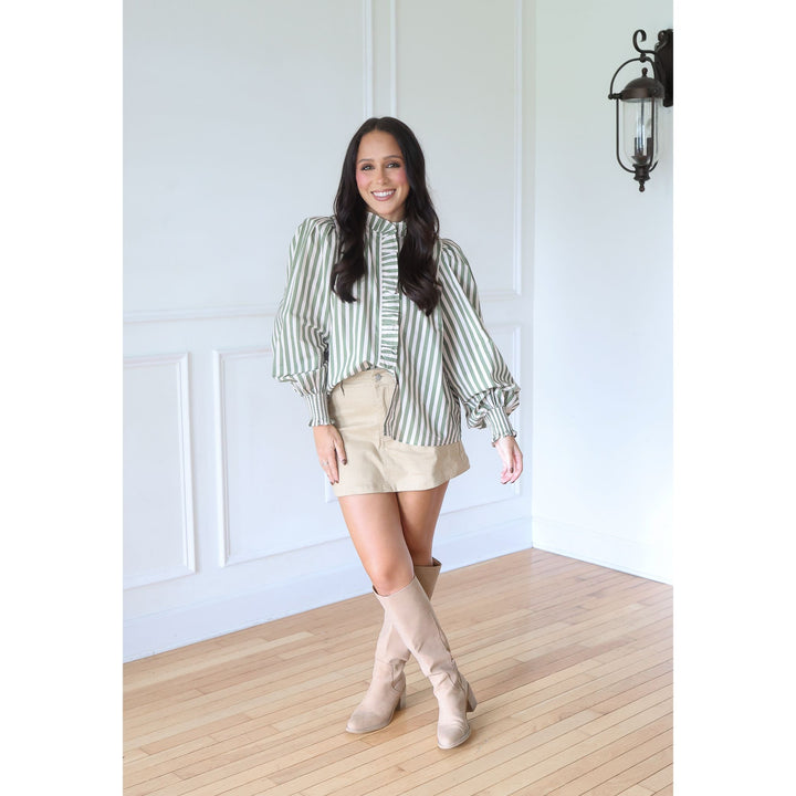 A woman modeling a green and white striped long-sleeve blouse with puffed balloon sleeves, paired with a beige mini skirt and beige knee-high boots, standing in a bright room with white paneled walls and light wood floors.