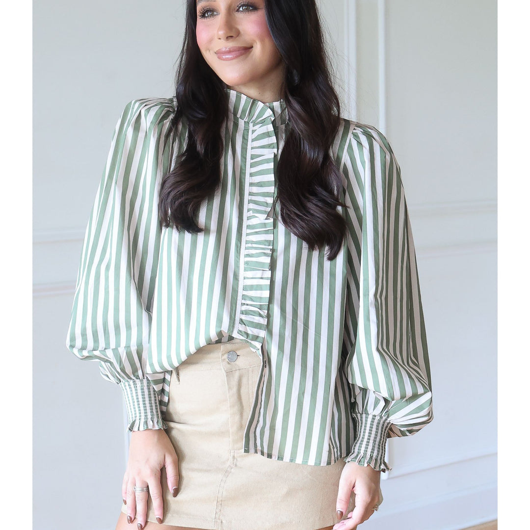 A woman wearing a green and white vertically striped blouse with ruffled trim and balloon sleeves, paired with a beige mini skirt, standing against a white paneled wall.