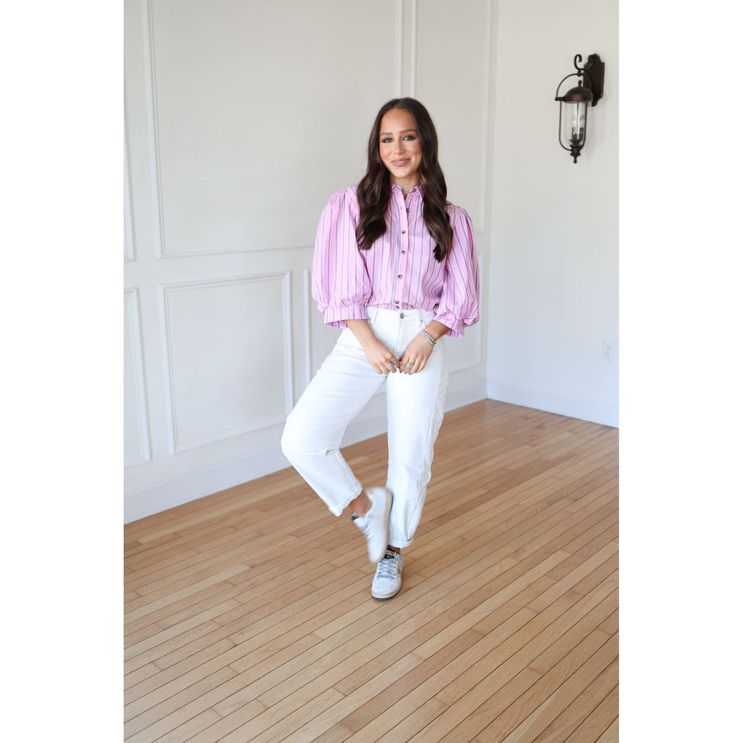 Woman wearing a pink striped shirt and white pants standing in a room with wooden flooring and white walls.