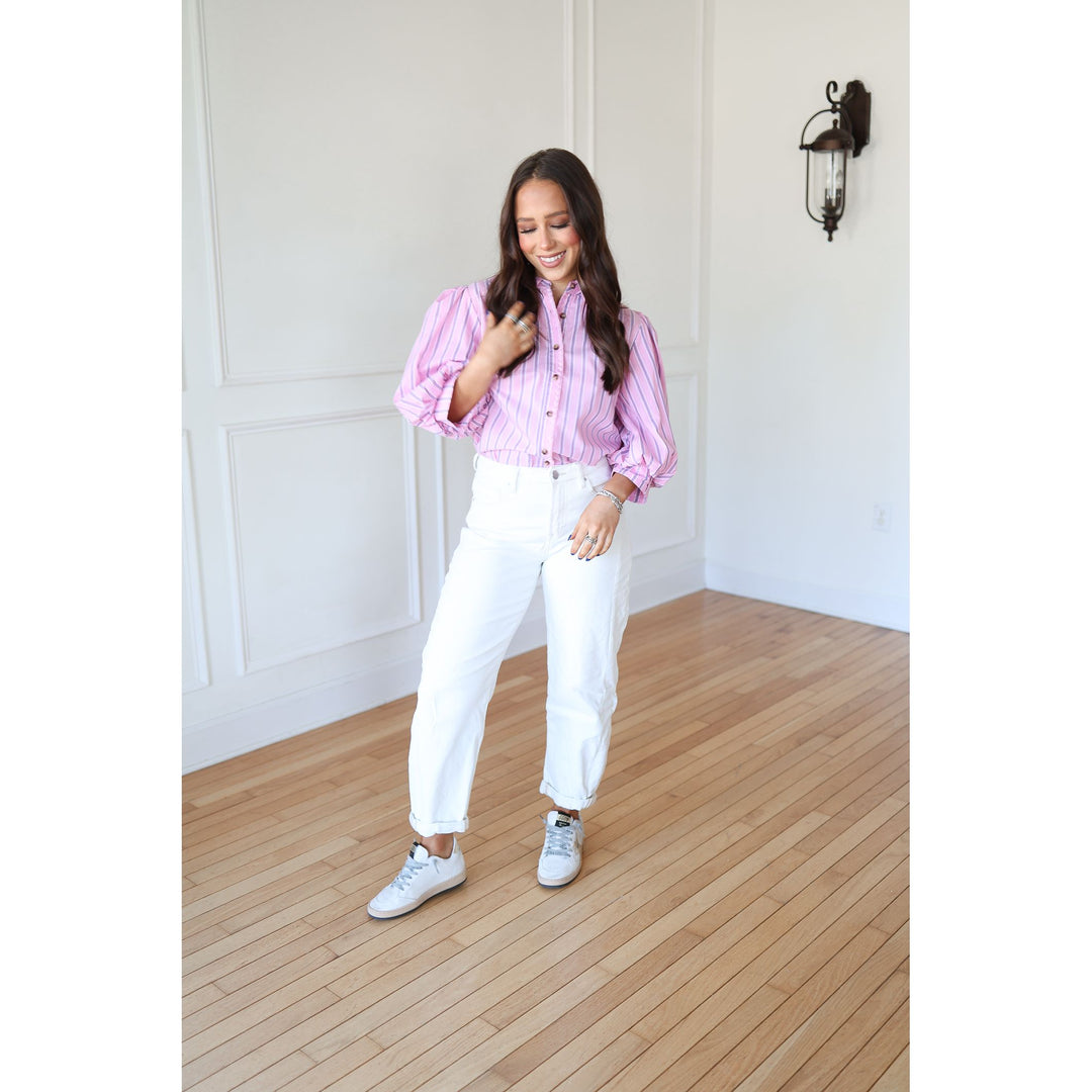 Woman wearing a pink striped shirt and white pants standing in a room with wooden flooring and white walls.