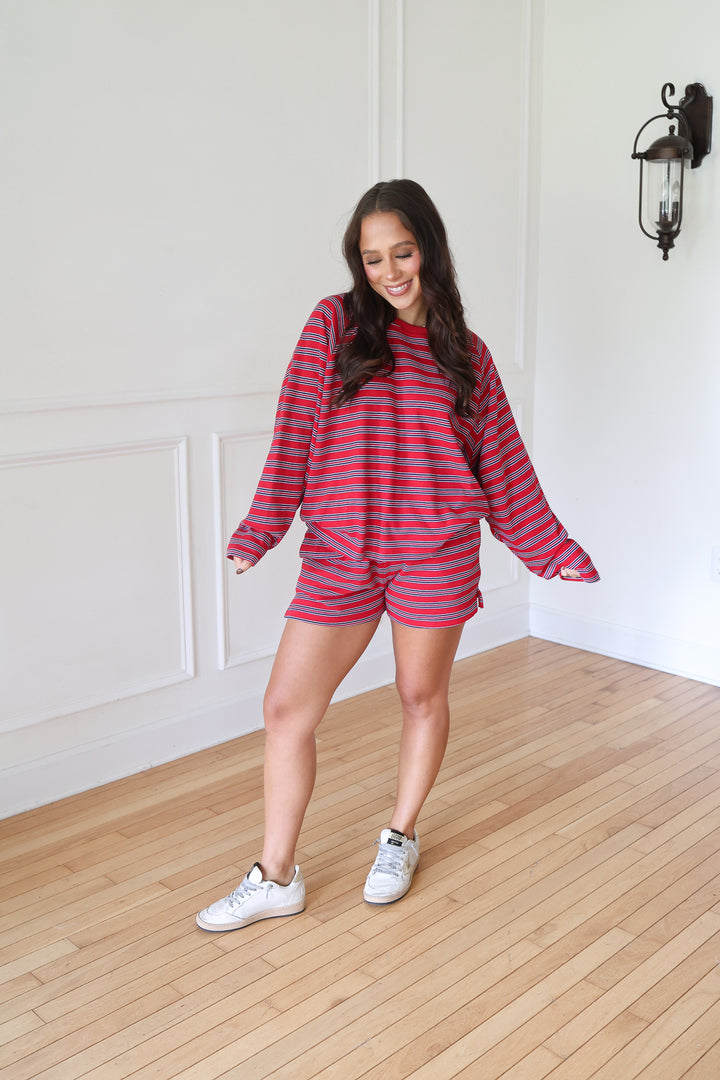 A woman modeling a matching red and navy striped lounge set consisting of a long-sleeve sweatshirt and shorts, paired with white sneakers,
