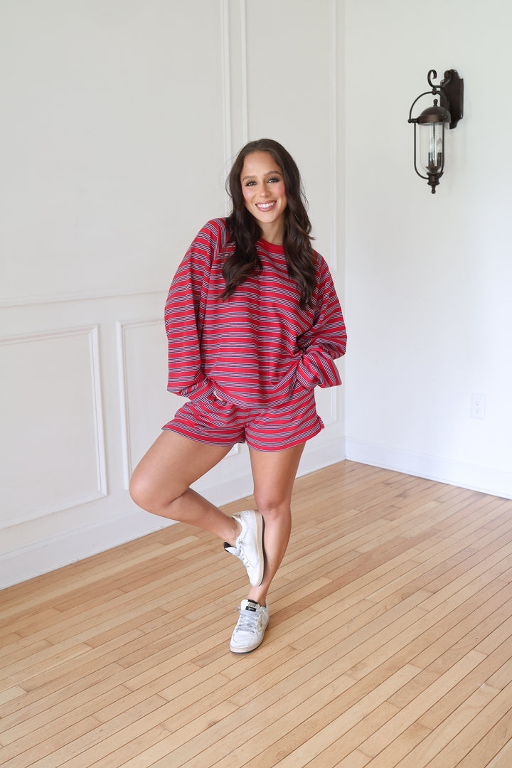 A woman posing in a matching red and navy striped lounge set, featuring a long-sleeve top and shorts, paired with white sneakers 