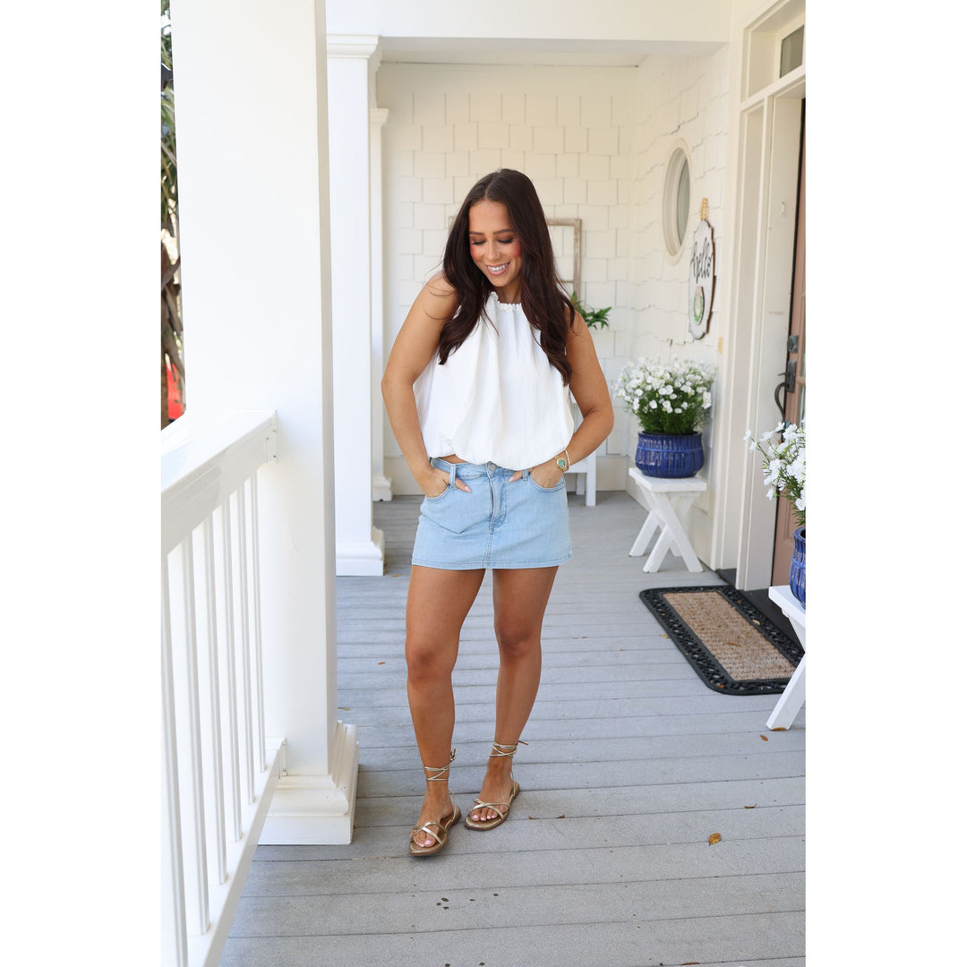 Woman standing on a porch wearing a white top and light blue shorts.