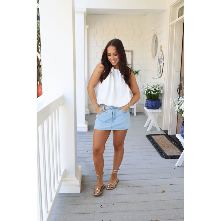 Woman standing on a porch wearing a white top and light blue shorts.