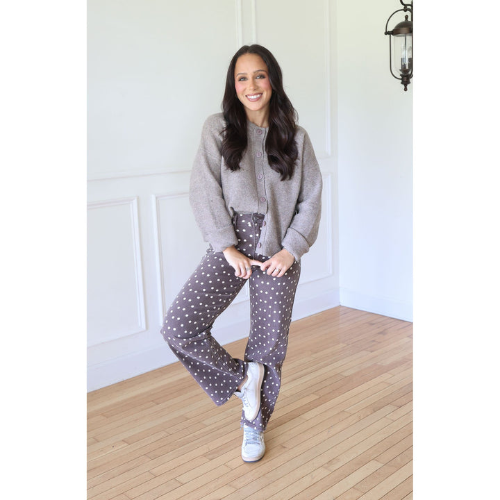 A woman posing in a cozy indoor setting with white paneled walls and light wood floors, wearing a light mocha knit cardigan, brown polka-dot pants, and white sneakers.