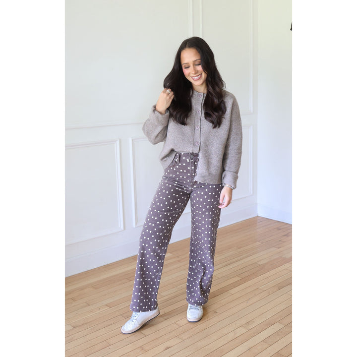A woman modeling a cozy outfit consisting of a mocha knit cardigan and high-waisted brown pants with white polka dots, paired with white sneakers, standing in a bright room with white paneled walls and light wood floors.