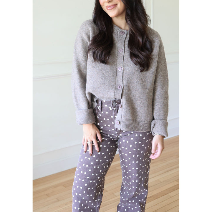 A woman wearing a mocha knit cardigan and brown pants with white polka dots, standing in a bright indoor room with light wood floors and white walls.