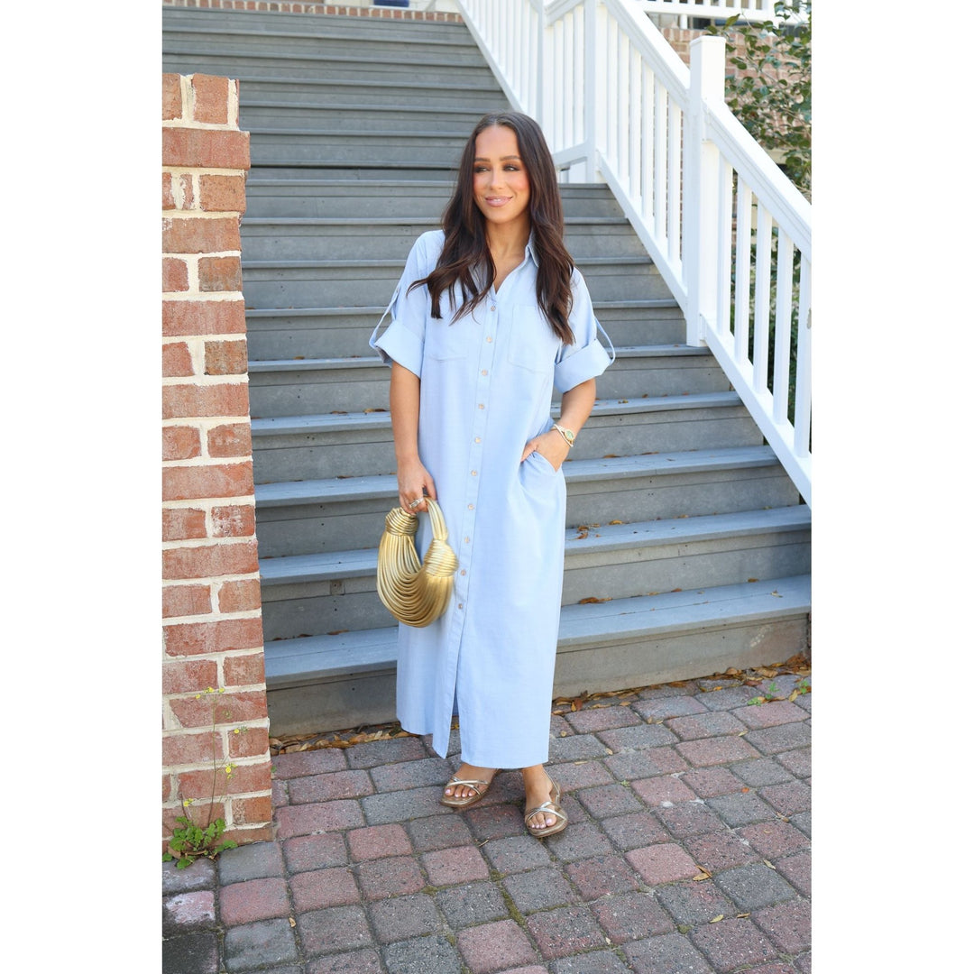 Woman in a light blue dress standing on a set of stairs with a brick wall and white railing in the background.