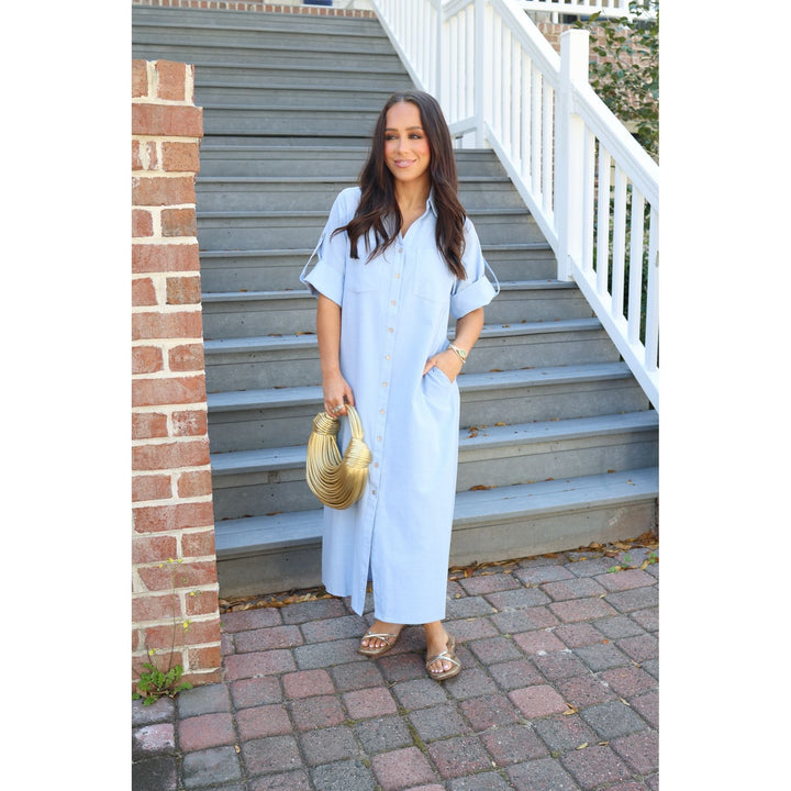Woman in a light blue dress standing on a set of stairs with a brick wall and white railing in the background.