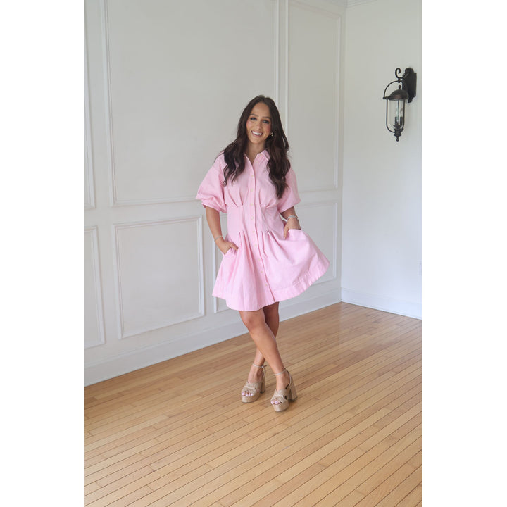 Woman in a pink dress standing in a room with wooden flooring and white walls.