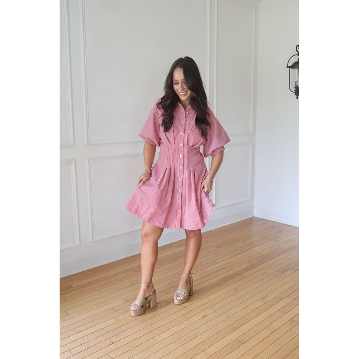Woman wearing a pink dress standing in a room with wooden flooring and white walls.