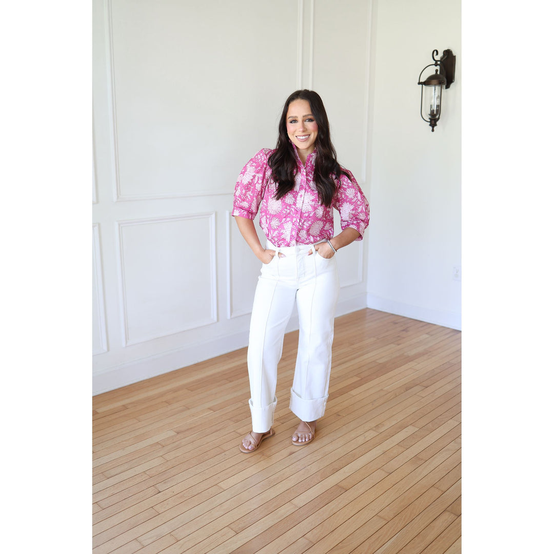 Woman in a pink floral blouse and white pants standing in a room with wooden flooring and white walls.