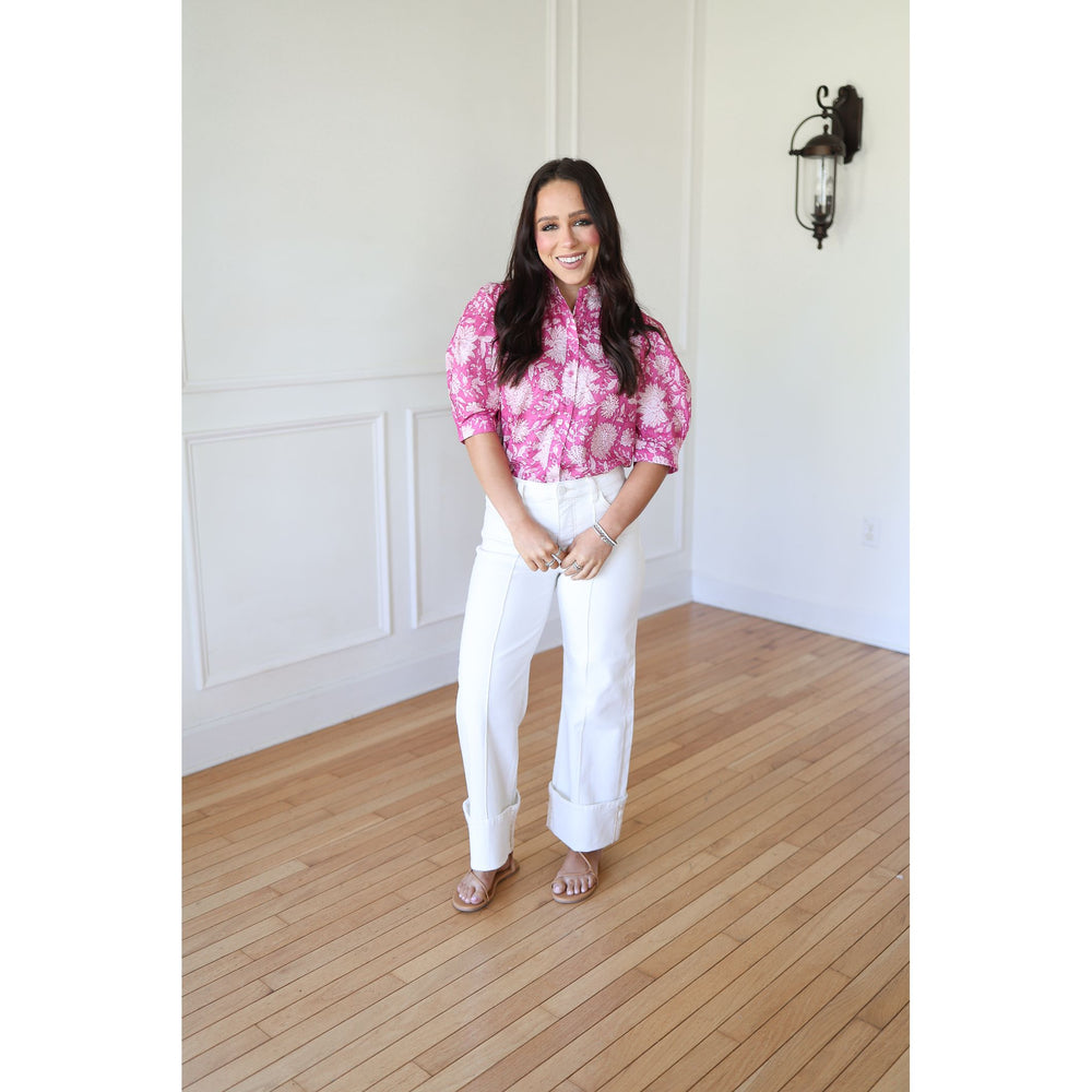 Woman wearing a pink floral blouse and white pants standing in a room with wooden flooring and white walls.