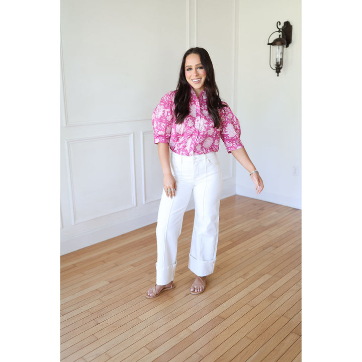 Woman wearing a pink floral blouse and white pants standing in a room with wooden flooring and white walls.