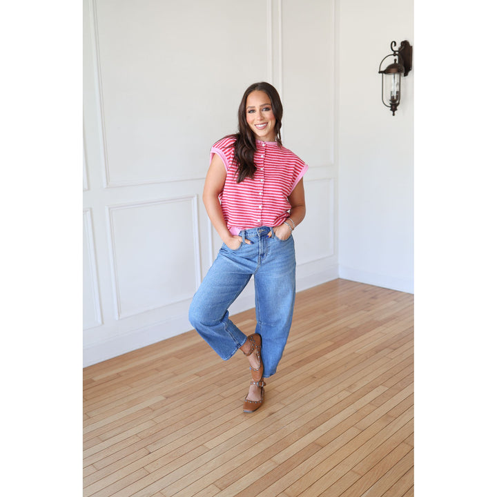 Woman wearing a pink striped shirt and blue jeans standing in a room with wooden flooring and white walls.
