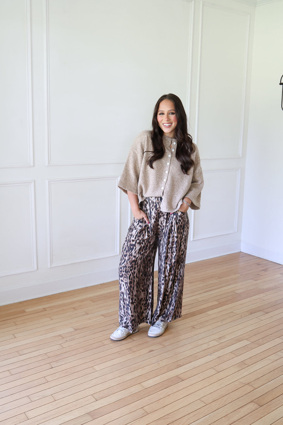 A woman stands in a bright room with white paneled walls and light wood floors, wearing a beige knit cardigan over a white top and wide-leg leopard-print pants with white sneakers.