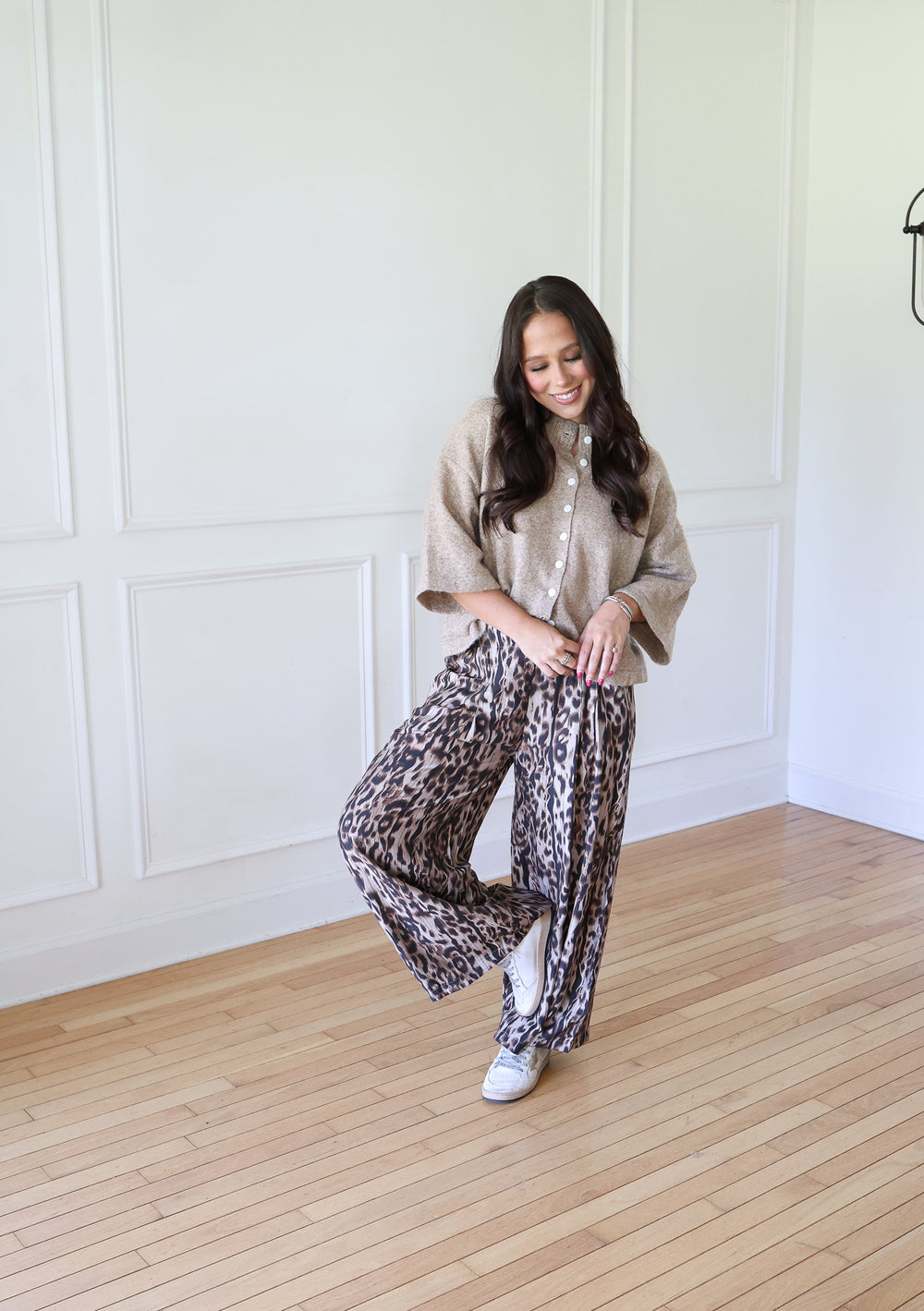 A woman posing in a bright room with white paneled walls and light wood floors, wearing a beige knit poncho over a white button-up shirt and wide-leg leopard-print pants.