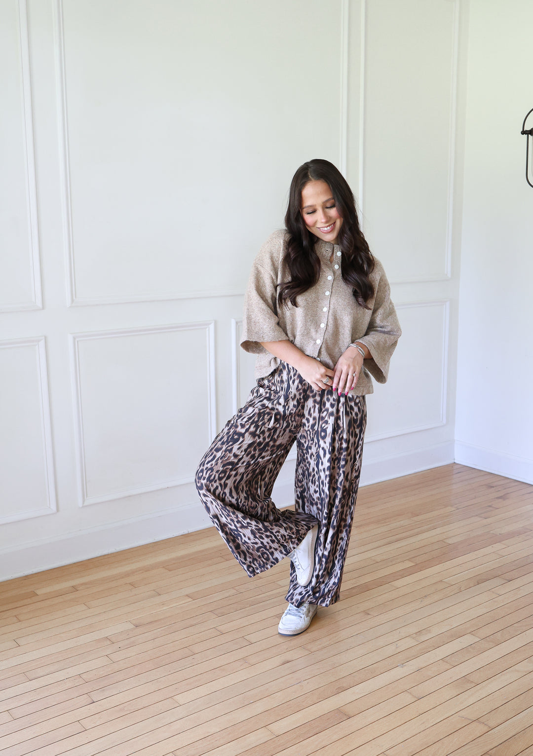 A woman posing in a bright room with white paneled walls and light wood floors, wearing a beige knit poncho over a white button-up shirt and wide-leg leopard-print pants.