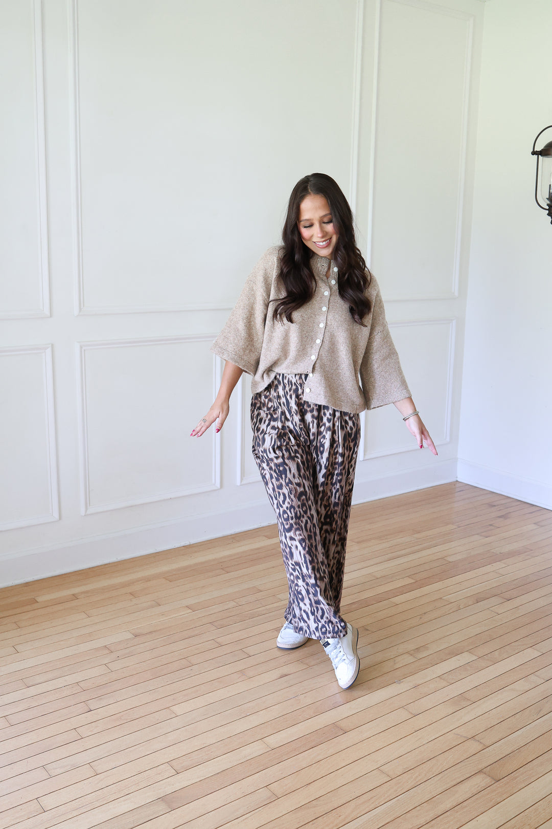 A woman modeling a beige knit cardigan and leopard-print pants in a bright room with white paneled walls and light wood floors.