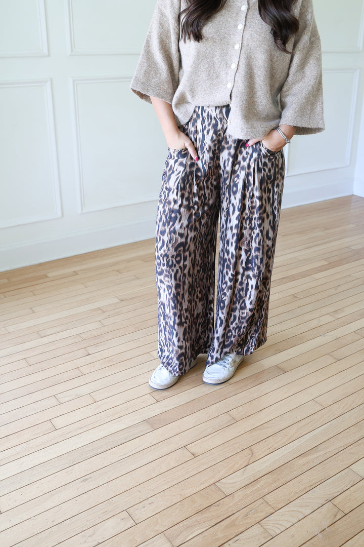 A woman modeling a beige knit cardigan and leopard-print pants in a bright room with white paneled walls and light wood floors.