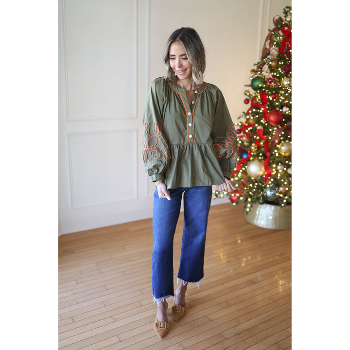 Woman in a green embroidered blouse and blue jeans standing in front of a decorated Christmas tree.