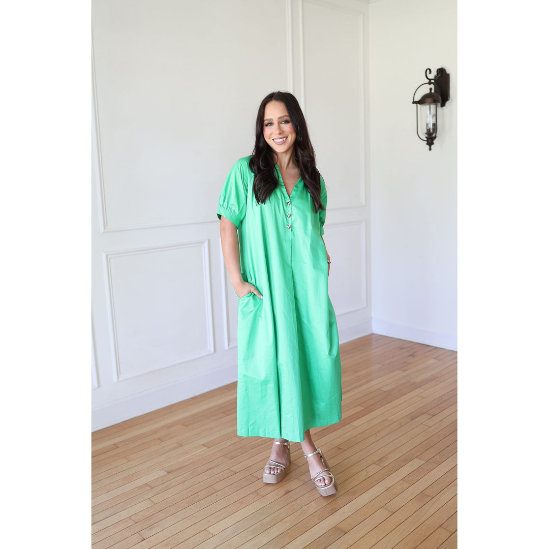 Woman wearing a green dress standing in a room with wooden flooring and white walls.