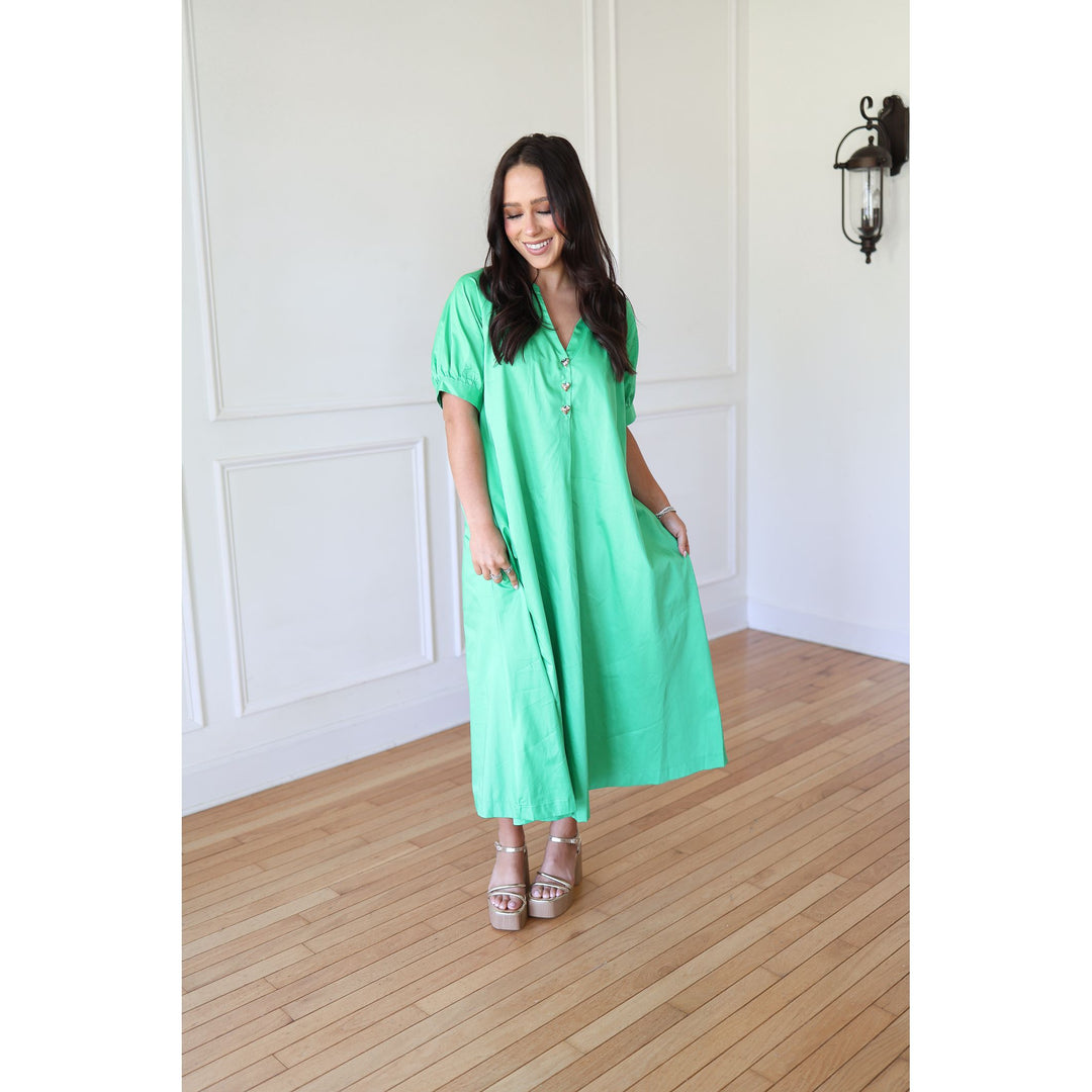 Woman wearing a green dress standing in a room with wooden flooring and white walls.