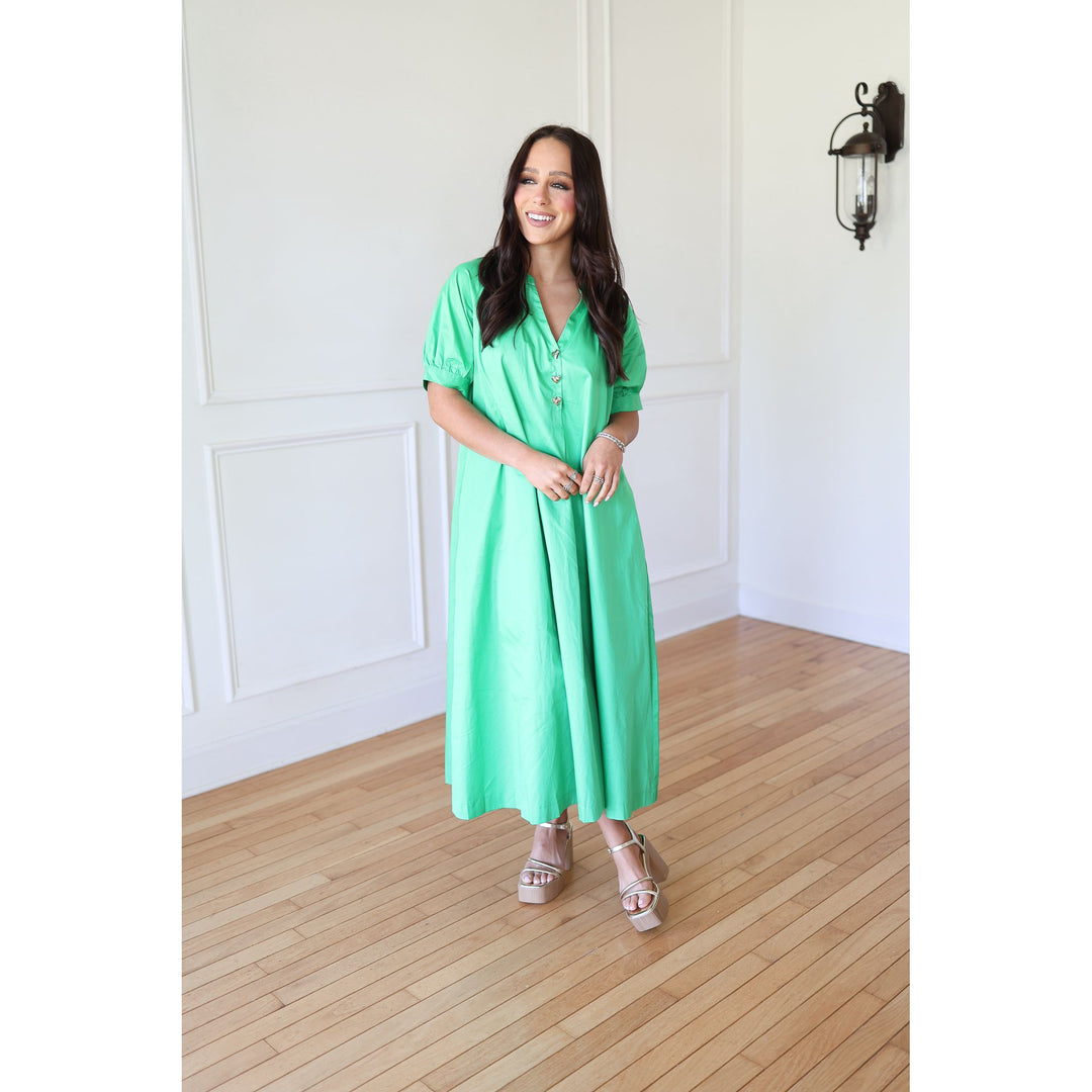 Woman wearing a green dress standing in a room with wooden flooring and white walls.