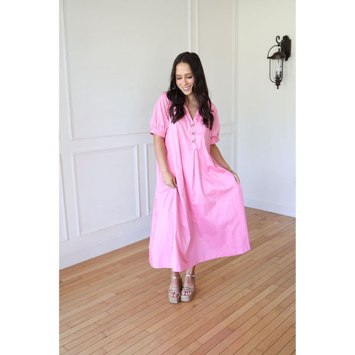 Woman wearing a pink dress standing in a room with wooden flooring and white walls.