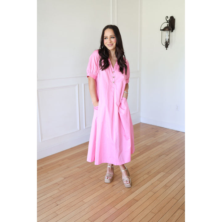 Woman wearing a pink dress standing in a room with wooden flooring and white walls.